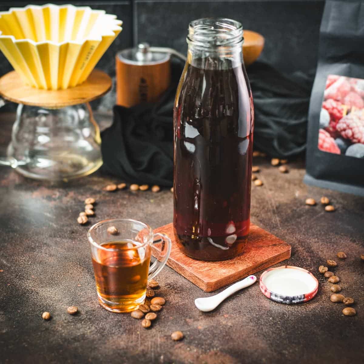 Amazing coffee syrup, in small glass next to a preserving bottle filled with coffee syrup, placed on brown ground next to coffee beans.