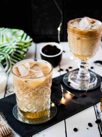Two gorgeous glasses filled with iced honey latte placed on black slate plate.