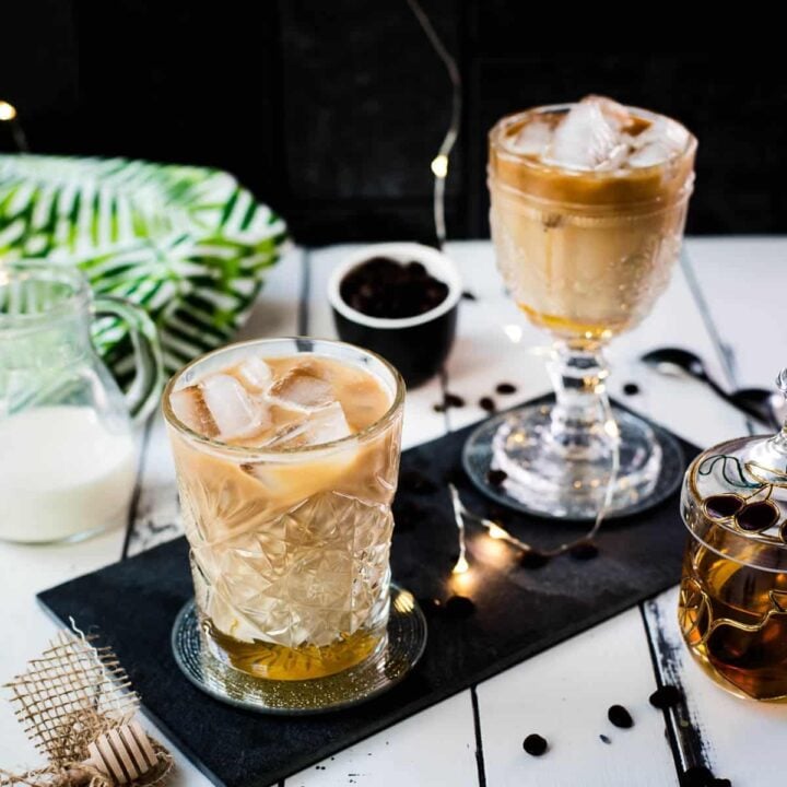 Two gorgeous glasses filled with iced honey latte placed on black slate plate.