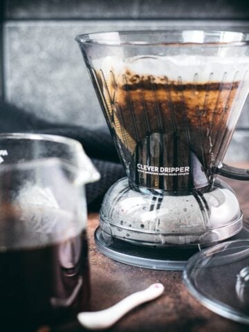 Filled clever dripper placed on dark ground, next to coffee filled glass carafe.