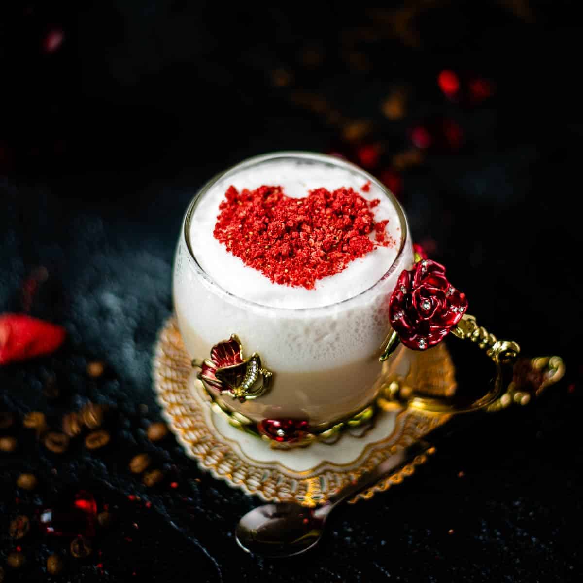 Gorgeous hot strawberry latte, served in a glass mug decorated with roses and butterflies. On top of the coffee is a red heart made from chopped freeze-dried hearts.