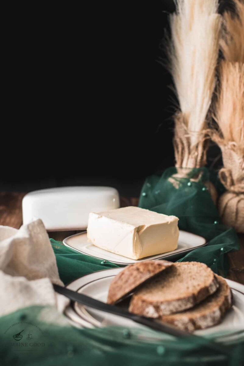 Cube of butter, placed on white plate with brown colored rim, next to a plate with sliced bread.