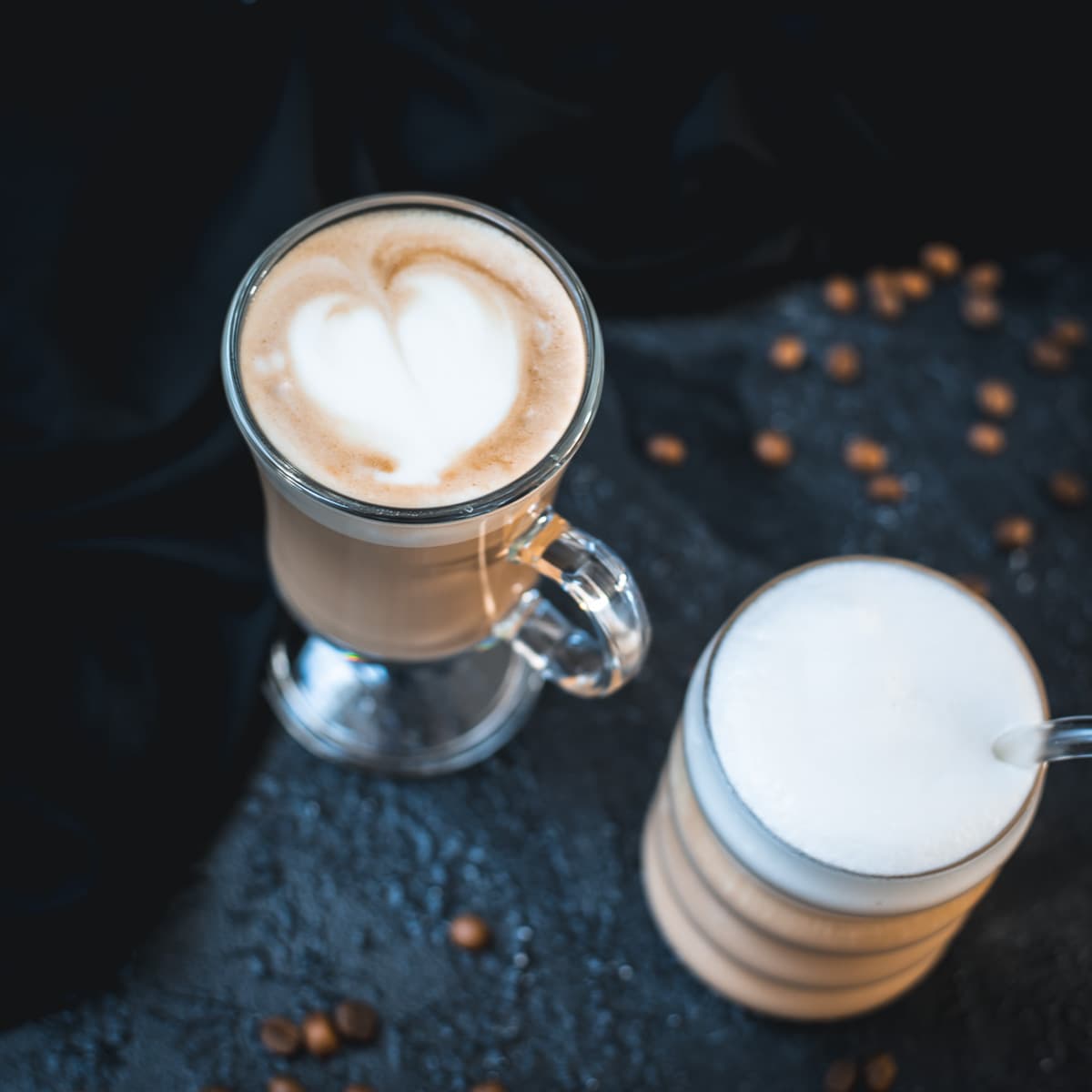 2 glasses filled with delicious vanilla latte. placed on dark ground. Left on has a latte art heart on top.