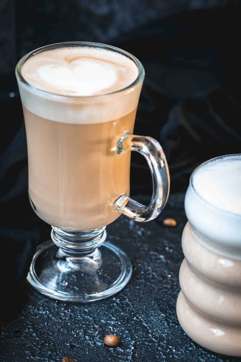 2 glasses filled with delicious vanilla latte. placed on dark ground. Left on has a latte art heart on top.