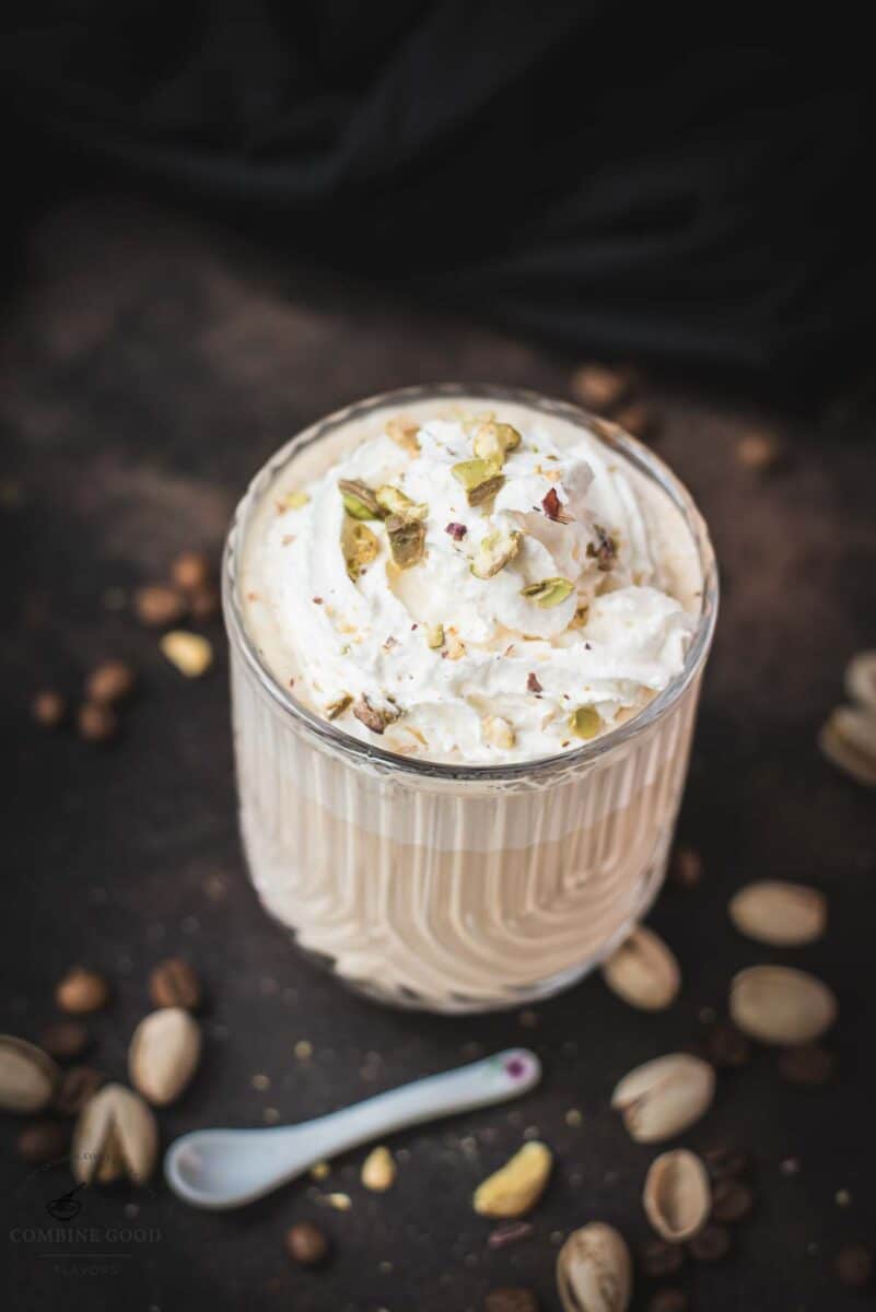 Divine pistachio latte with pistachio liqueur, served in beautiful drinking glass, resting on dark surface scattered with coffee beans and pistachios.