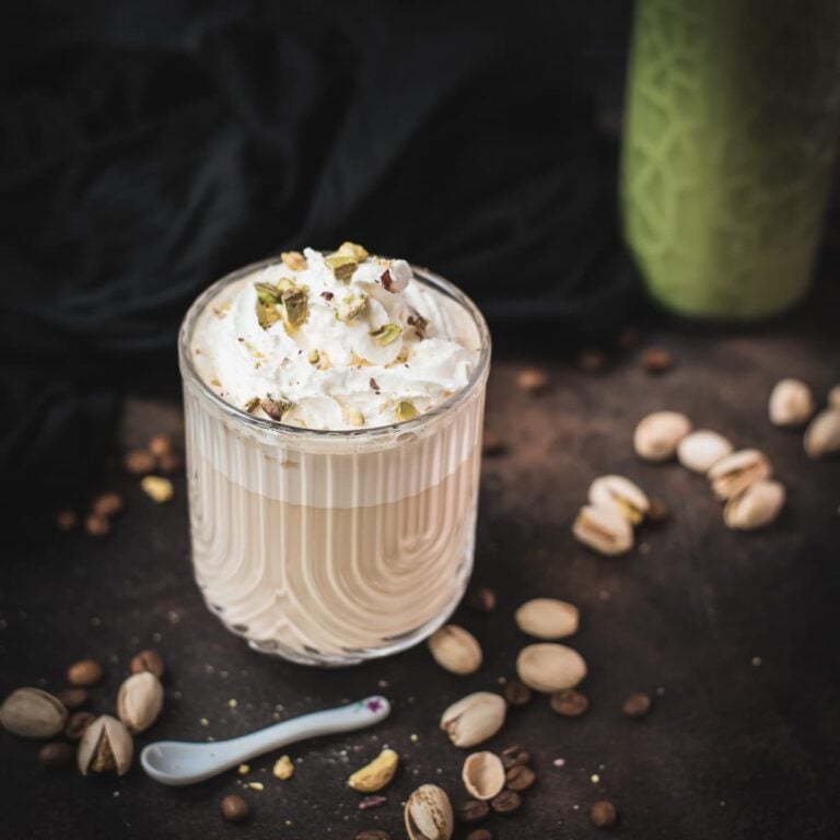 Divine pistachio latte with pistachio liqueur, served in beautiful drinking glass, resting on dark surface scattered with coffee beans and pistachios.