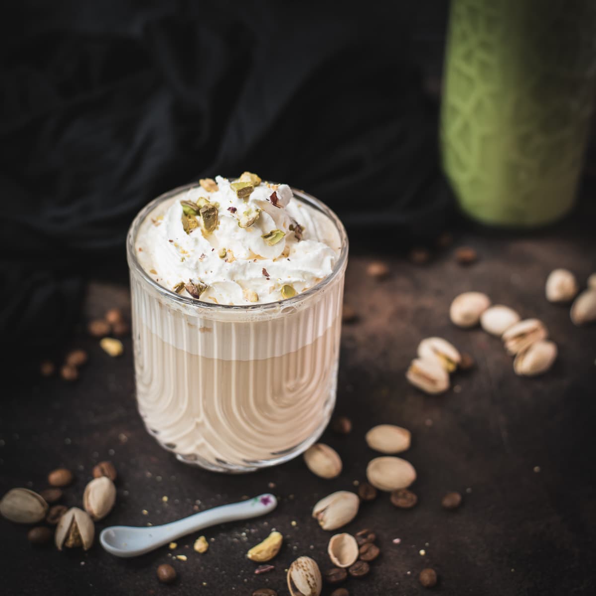 Divine pistachio latte with pistachio liqueur, served in beautiful drinking glass, resting on dark surface scattered with coffee beans and pistachios.