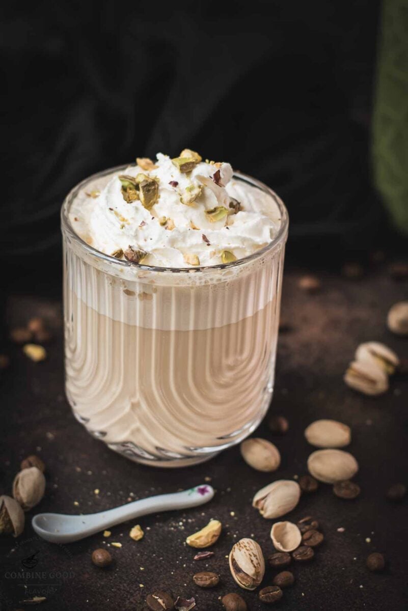 Divine pistachio latte with pistachio liqueur, served in beautiful drinking glass, resting on dark surface scattered with coffee beans and pistachios.