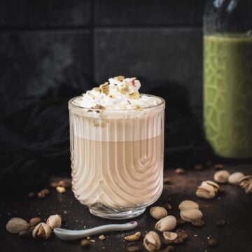 Divine pistachio latte with pistachio liqueur, served in beautiful drinking glass, resting on dark surface scattered with coffee beans and pistachios.