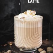 Divine pistachio latte with pistachio liqueur, served in beautiful drinking glass, resting on dark surface scattered with coffee beans and pistachios.