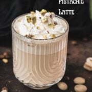 Divine pistachio latte with pistachio liqueur, served in beautiful drinking glass, resting on dark surface scattered with coffee beans and pistachios.