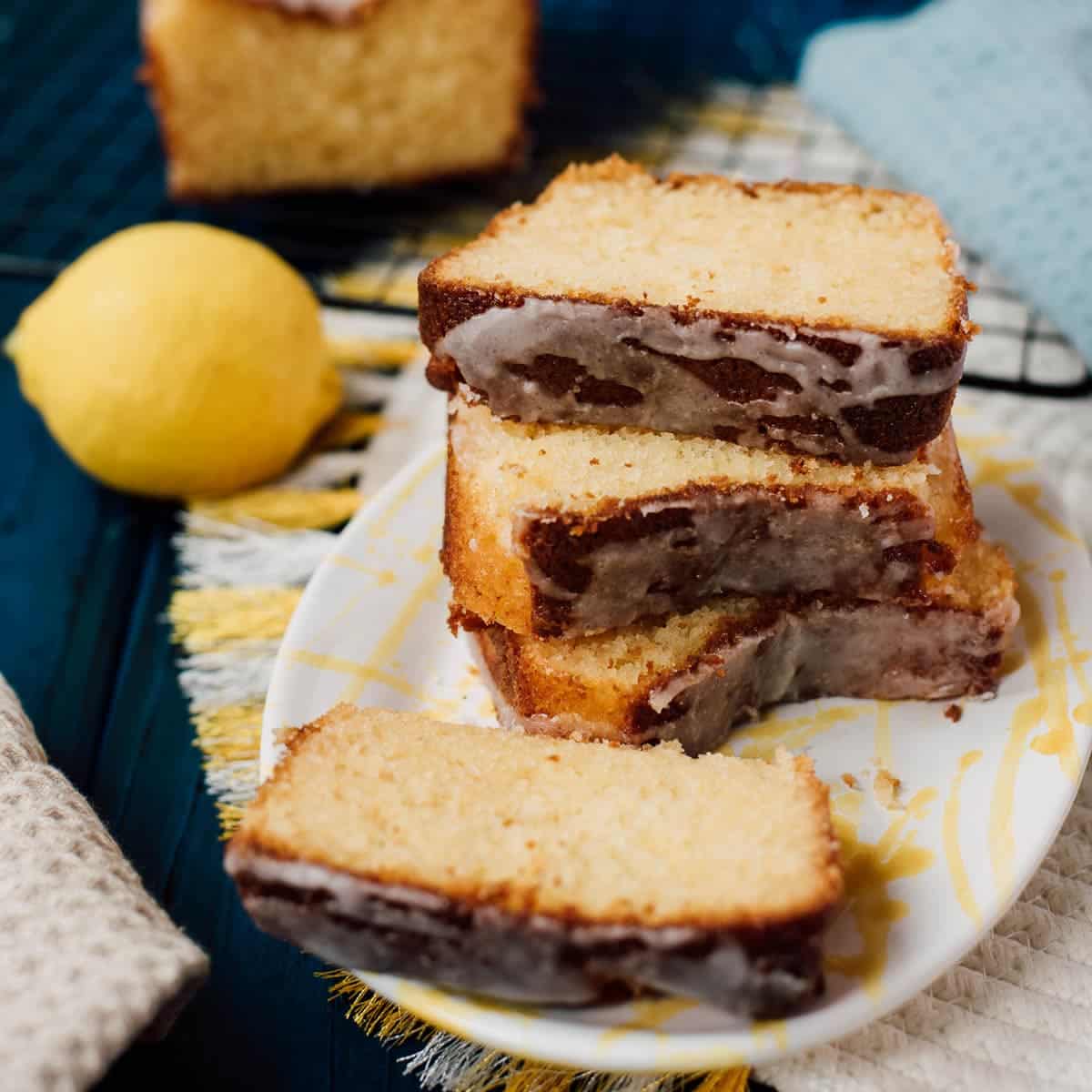 Sliced and stacked lemon loaf cake slices, placed on white and yellow plate, next to a lemon.
