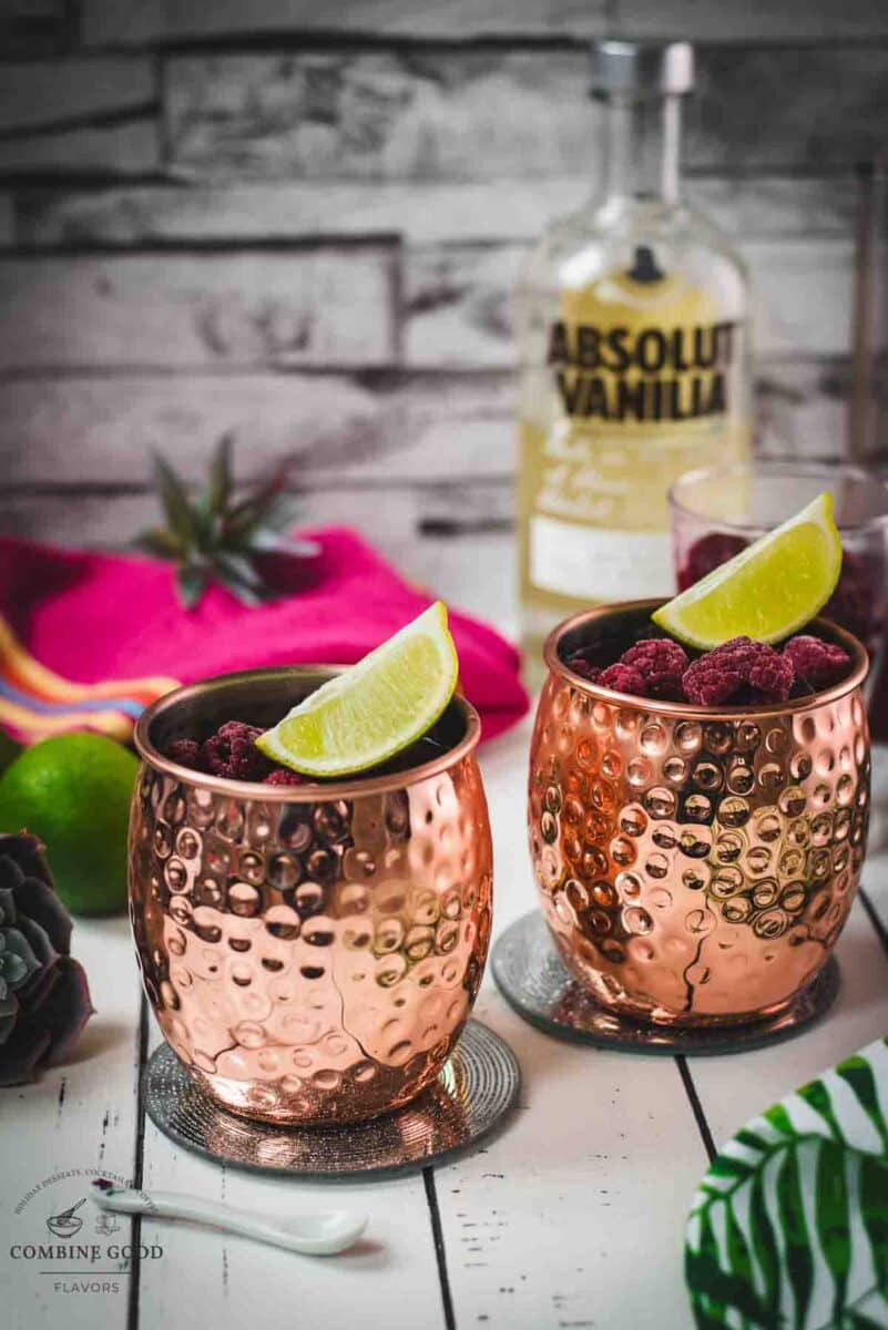 Two icy copper mugs holding a raspberry mule, decorated with lime wedges and frozen raspberries on a white table.