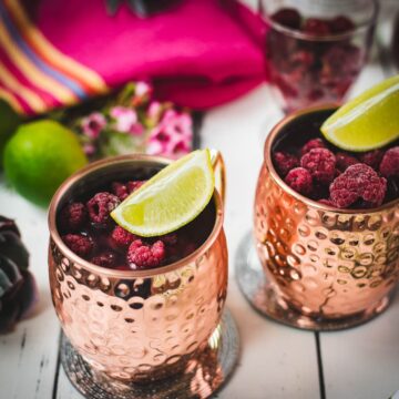 Two icy copper mugs holding a raspberry mule, decorated with lime wedges and frozen raspberries on a white table.