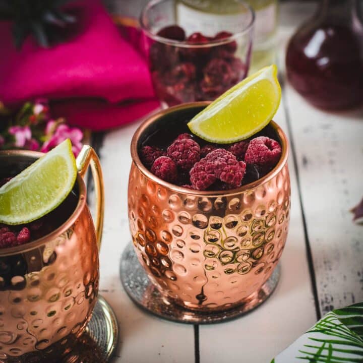 Two icy copper mugs holding a raspberry mule, decorated with lime wedges and frozen raspberries on a white table.