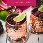 Two icy copper mugs holding a raspberry mule, decorated with lime wedges and frozen raspberries on a white table.