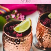 Two icy copper mugs holding a raspberry mule, decorated with lime wedges and frozen raspberries on a white table.