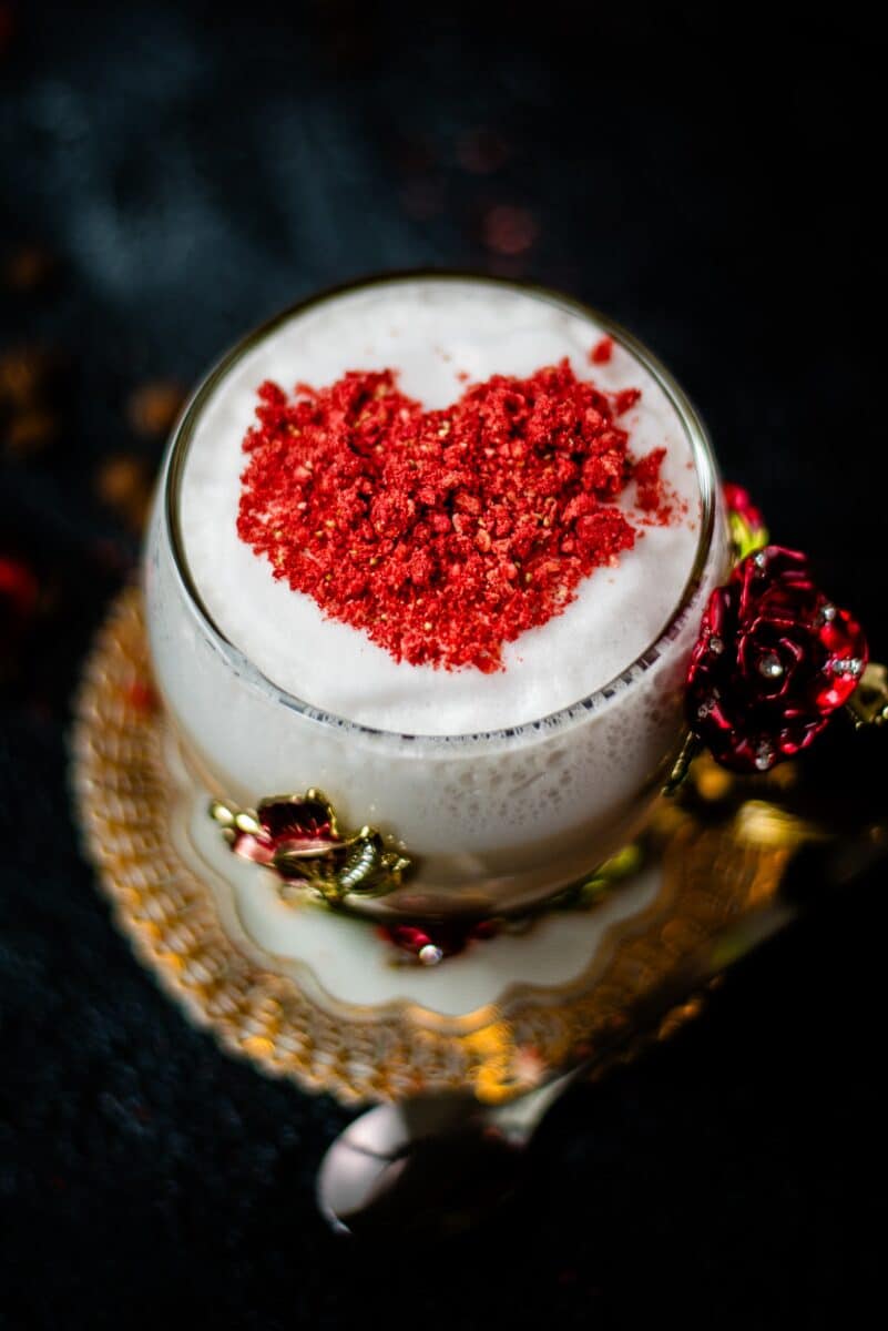 Gorgeous hot strawberry latte, served in a glass mug decorated with roses and butterflies. On top of the coffee is a red heart made from chopped freeze-dried hearts.