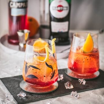 Gorgeous Americano cocktails. One garnished with a lemon twist the other with an orange wedge. Both glasses placed together on a black slate plate.