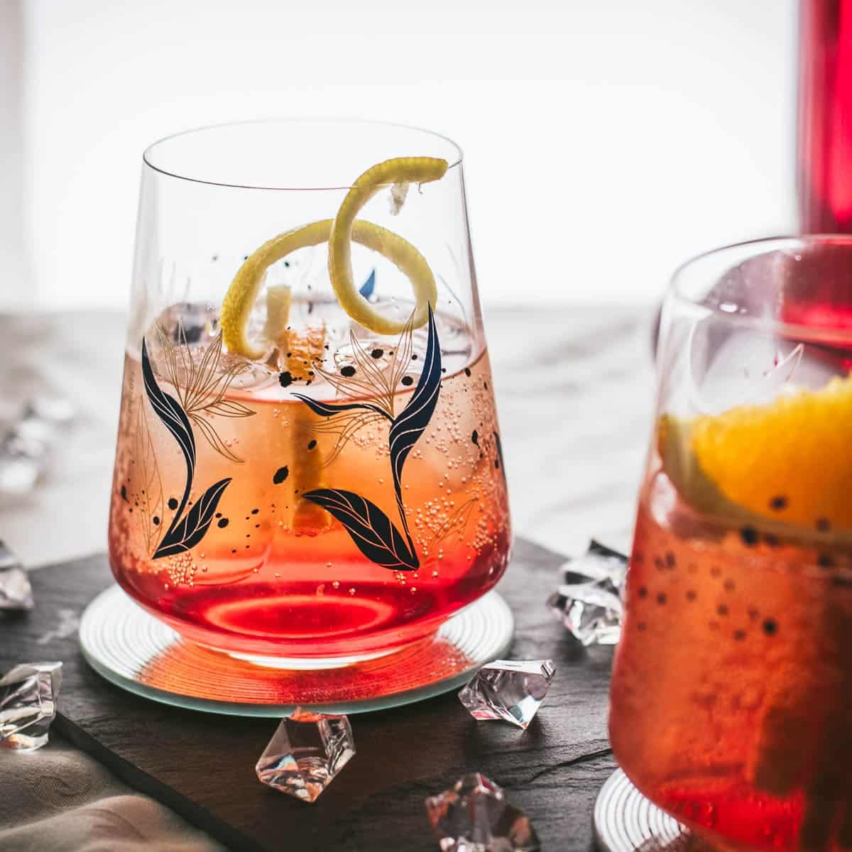 Gorgeous Americano cocktails. One garnished with a lemon twist the other with an orange wedge. Both glasses placed together on a black slate plate.