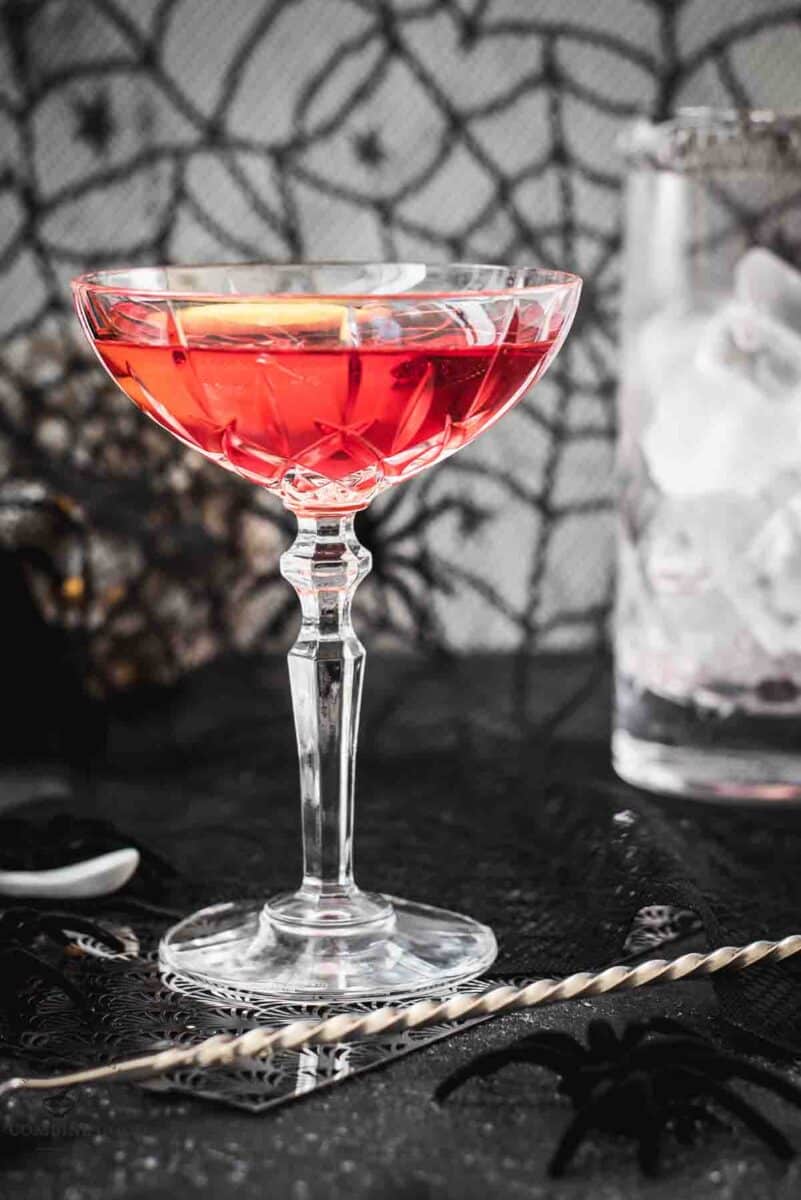 Cocktail Glass filled with a stunning red old pal cocktail, garnished with a lemon zest, and placed on spider web covered ground.