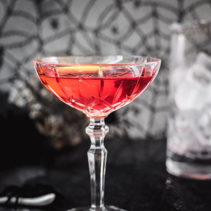Cocktail Glass filled with a stunning red old pal cocktail, garnished with a lemon zest, and placed on spider web covered ground.
