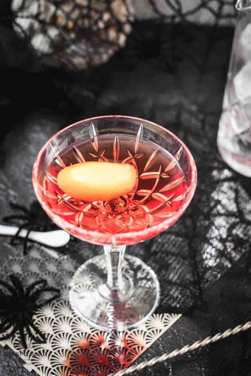 Cocktail Glass filled with a stunning red old pal cocktail, garnished with a lemon zest, and placed on spider web covered ground.