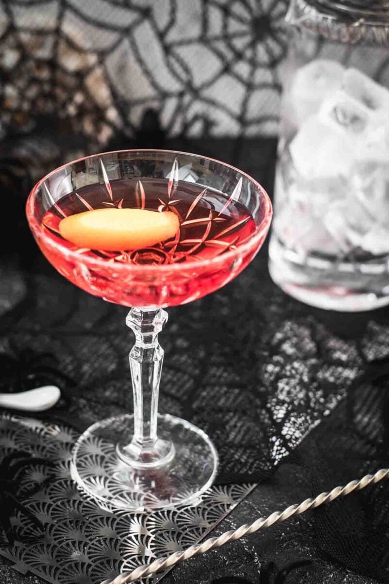 Cocktail Glass filled with a stunning red old pal cocktail, garnished with a lemon zest, and placed on spider web covered ground.