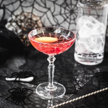 Cocktail Glass filled with a stunning red old pal cocktail, garnished with a lemon zest, and placed on spider web covered ground.
