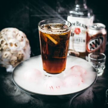 Halloween bourbon and root beer cocktail surrounded by smoke, dragon eyes, skulls, and spiderwebs.