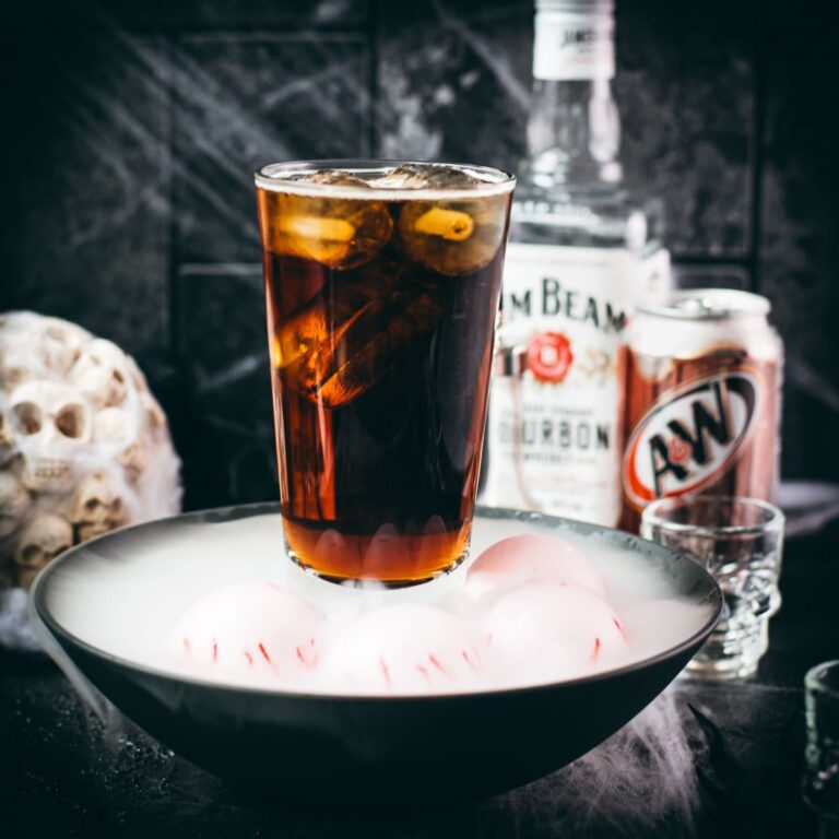 Dark root beer cocktail in a smoky Halloween setting with dragon eyes and skull decorations.