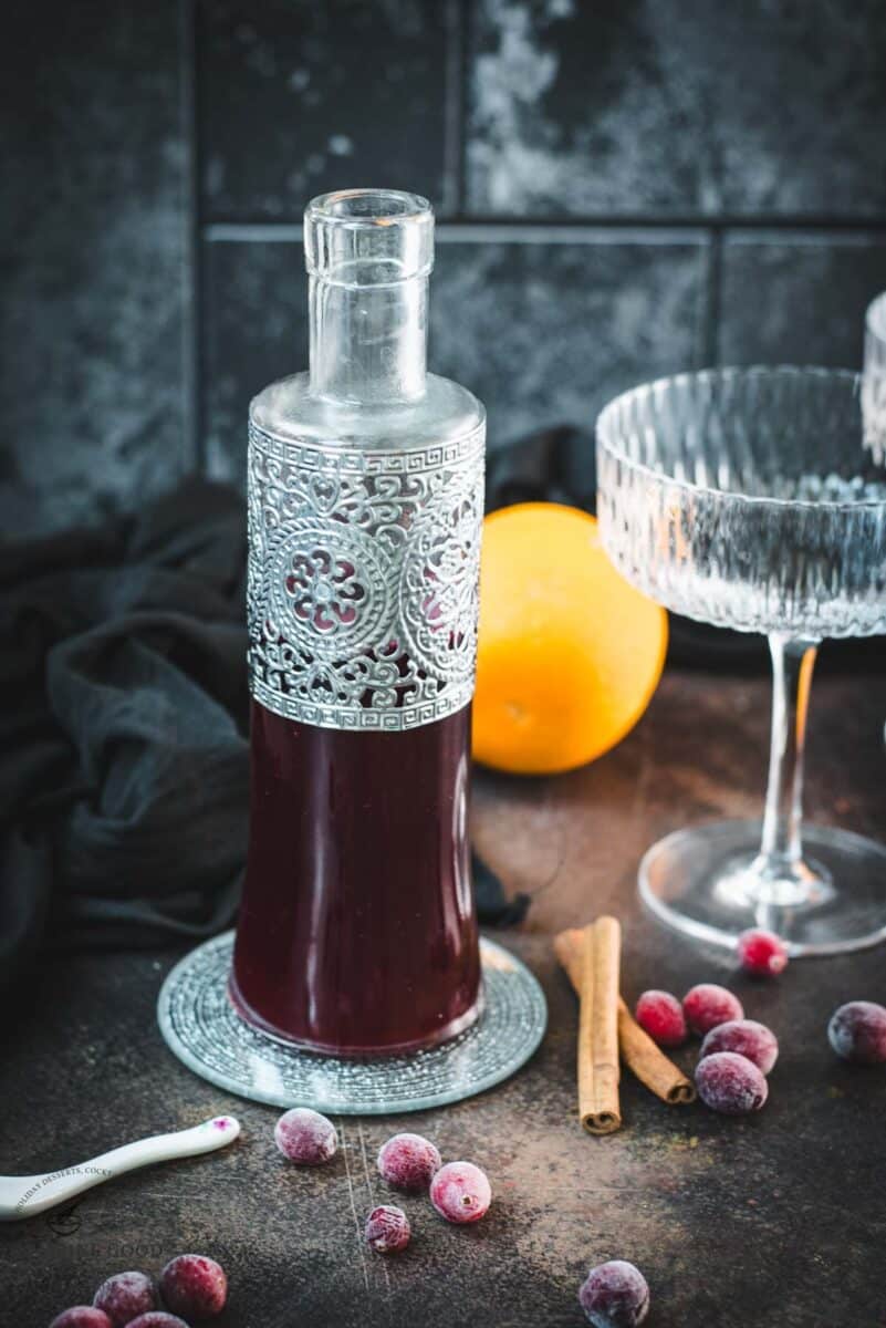 Cranberry simple syrup bottle surrounded by frozen cranberries and cinnamon sticks on a dark surface