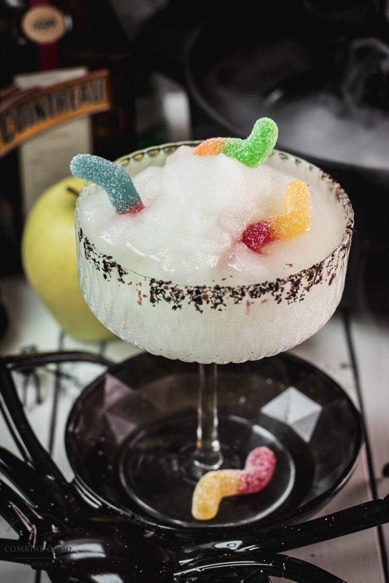 Close-up of gummy worms peeking from the vivid green frozen cocktail beside the coarse black sugar rim.