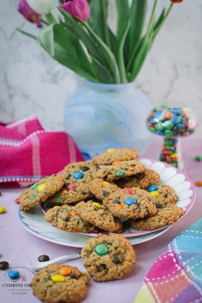 Oatmeal M&M chocolate chip cookies on a plate with colorful M&M’s placed in a mushroom-shaped glass beside spring tulips.