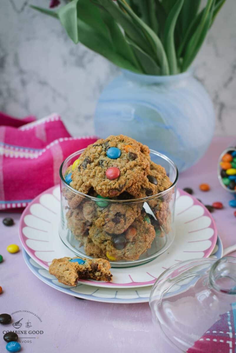 Oatmeal M&M cookies in a glass cookie jar with a partially eaten cookie in front and scattered M&M’s.
