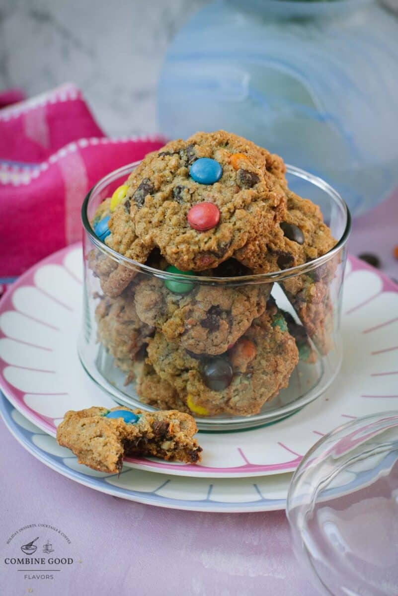 Oatmeal M&M cookies in a glass cookie jar with a partially eaten cookie in front and scattered M&M’s.
