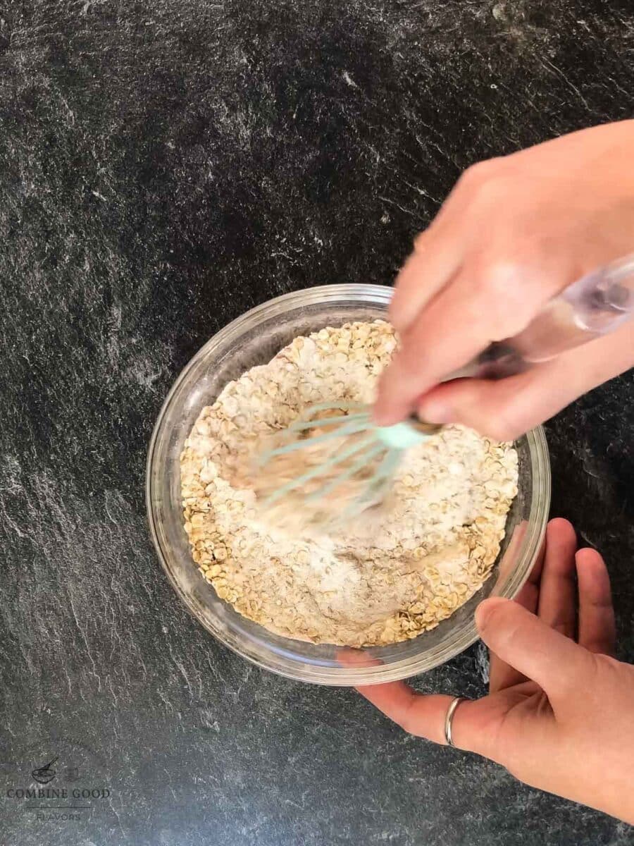 Flour, baking soda, salt, cinnamon, and rolled oats getting whisked in a mixing bowl.