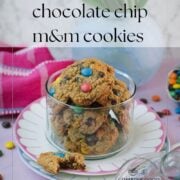 Oatmeal M&M cookies in a glass cookie jar with a partially eaten cookie in front and scattered M&M’s.