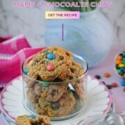 Oatmeal M&M cookies in a glass cookie jar with a partially eaten cookie in front and scattered M&M’s.