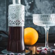 Homemade cranberry simple syrup in a decorative glass bottle with frozen cranberries, cinnamon, and orange