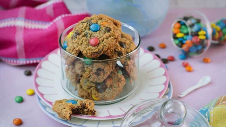 Glass cookie jar filled with oatmeal M&M cookies on pink and light blue plates with colorful M&M&rsquo;s scattered nearby.