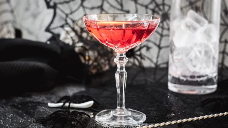 Cocktail Glass filled with a stunning red old pal cocktail, garnished with a lemon zest, and placed on spider web covered ground.