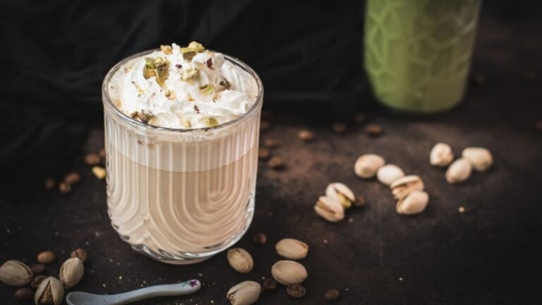 Divine pistachio latte with pistachio liqueur, served in beautiful drinking glass, resting on dark surface scattered with coffee beans and pistachios.