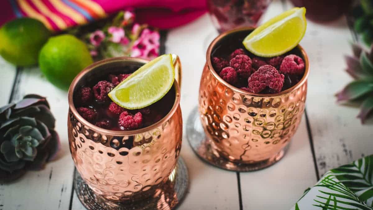 Two icy copper mugs holding a raspberry mule, decorated with lime wedges and frozen raspberries on a white table.